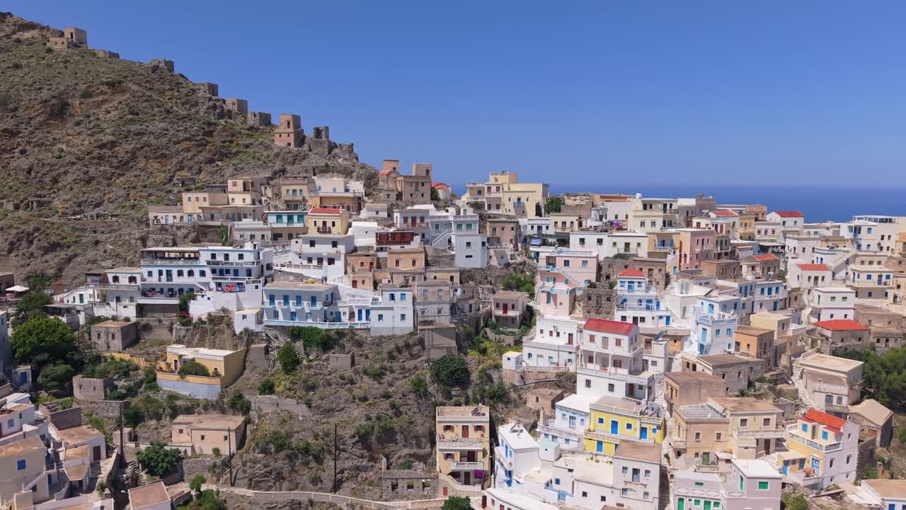Drone pull-back movement reveals the traditional village of Olympos in Karpathos, showcasing its hillside location, colorful houses, and the stunning surrounding Aegean landscape