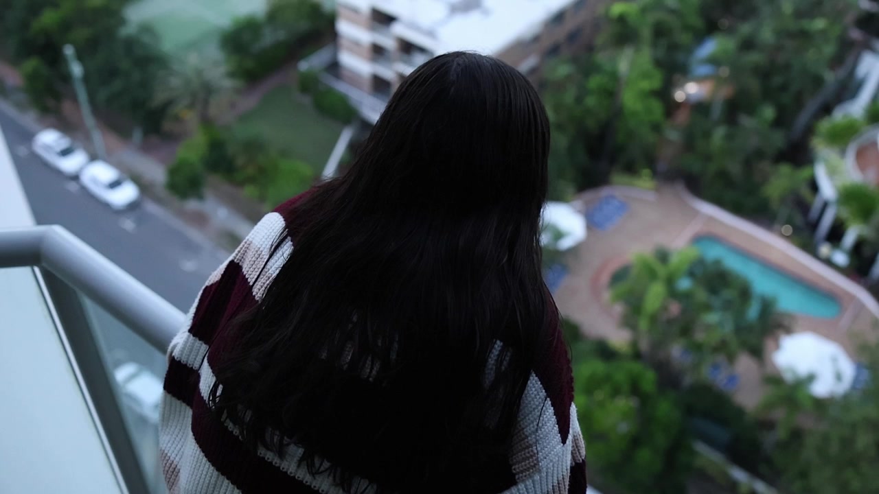 A woman with long dark hair, wrapped in a striped blanket, gazes thoughtfully from a high-rise balcony at a city street and swimming pool below, shot in soft daylight.
