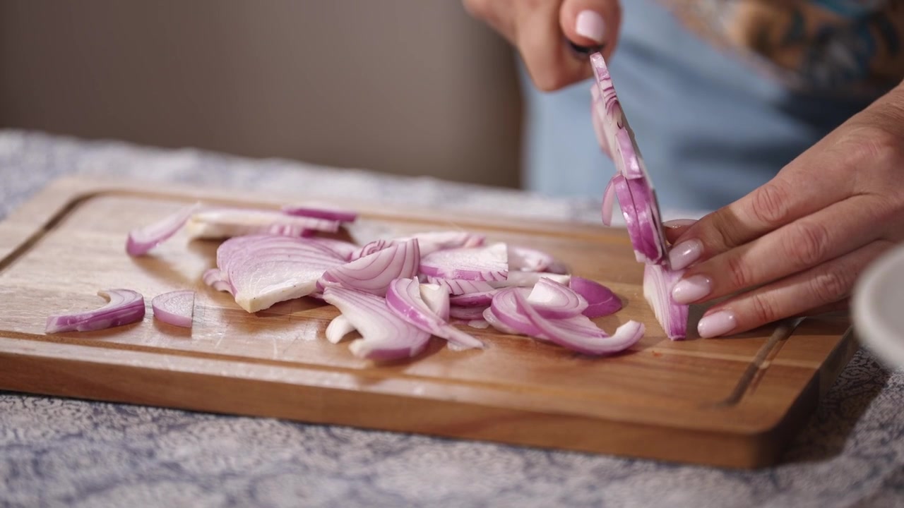 Primo piano di mani che affettano cipolla rossa fresca con un coltello da cucina su un tagliere di legno. Preparazione del cibo per la cucina casalinga con ingredienti freschi in una cucina rustica