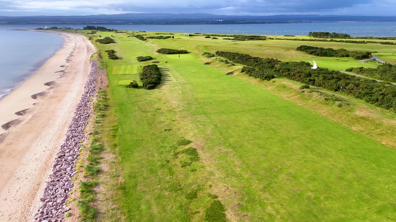 Drone camera glides above a seaside golf course in Rosemarkie, Scotland, revealing golfers, sandy beach, grassy fairways, and dramatic coastal scenery in bright daylight