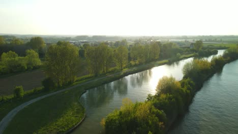 Vista-Aérea-De-La-Puesta-De-Sol-Sobre-Un-Río-Que-Revela-El-Pueblo-De-Clairmarais,-Francia-En-La-Distancia