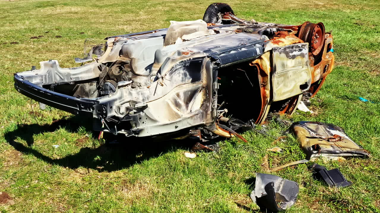 Burned car frame on grass field showing fire damage and stripped interior remains