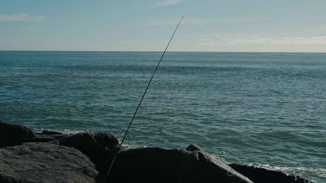 Canna da pesca angolata su una costa rocciosa con un oceano blu calmo sotto un cielo sereno.