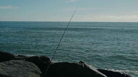 fishing rod angled over rocky shoreline with calm blue ocean under clear sky