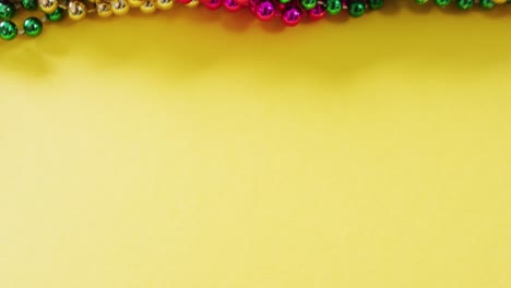 Video-of-pink,-gold-and-green-mardi-gras-carnival-beads-on-yellow-background-with-copy-space