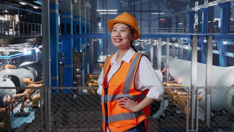 side view of asian female engineer with safety helmet in factory manufacture of wind turbines. standing with arms akimbo looking around, checking