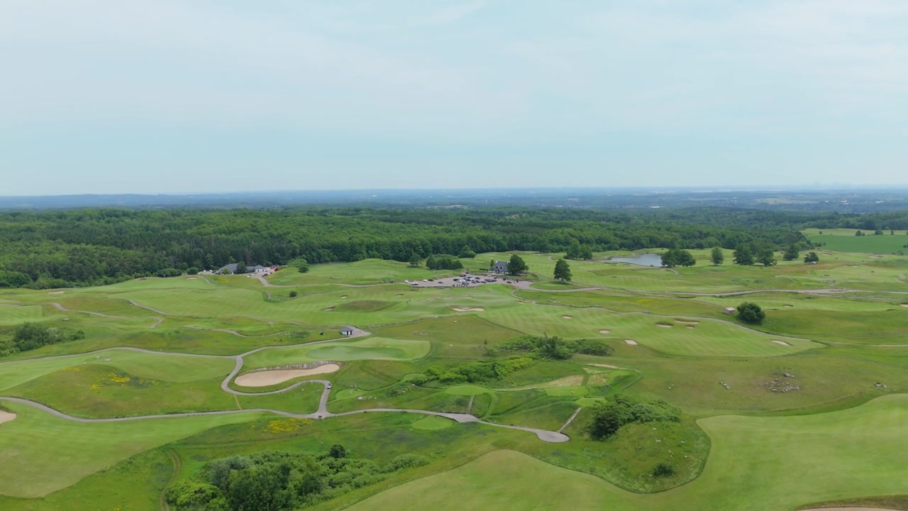 Sweeping aerial footage captures the scenic expanse of Devil's Paintbrush Golf Course near Toronto, with rolling fairways, sand bunkers, lush greens, winding paths, and woodlands under soft daylight.