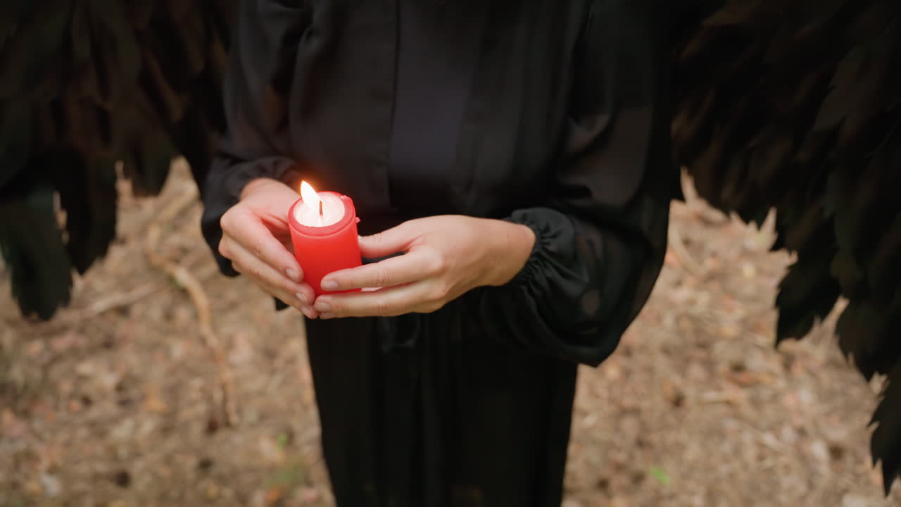Close up of woman dressed in black with large black wings holding burning red candle in forest, performing quiet ritual symbolizing faith, energy, devotion, and mystical connection with nature