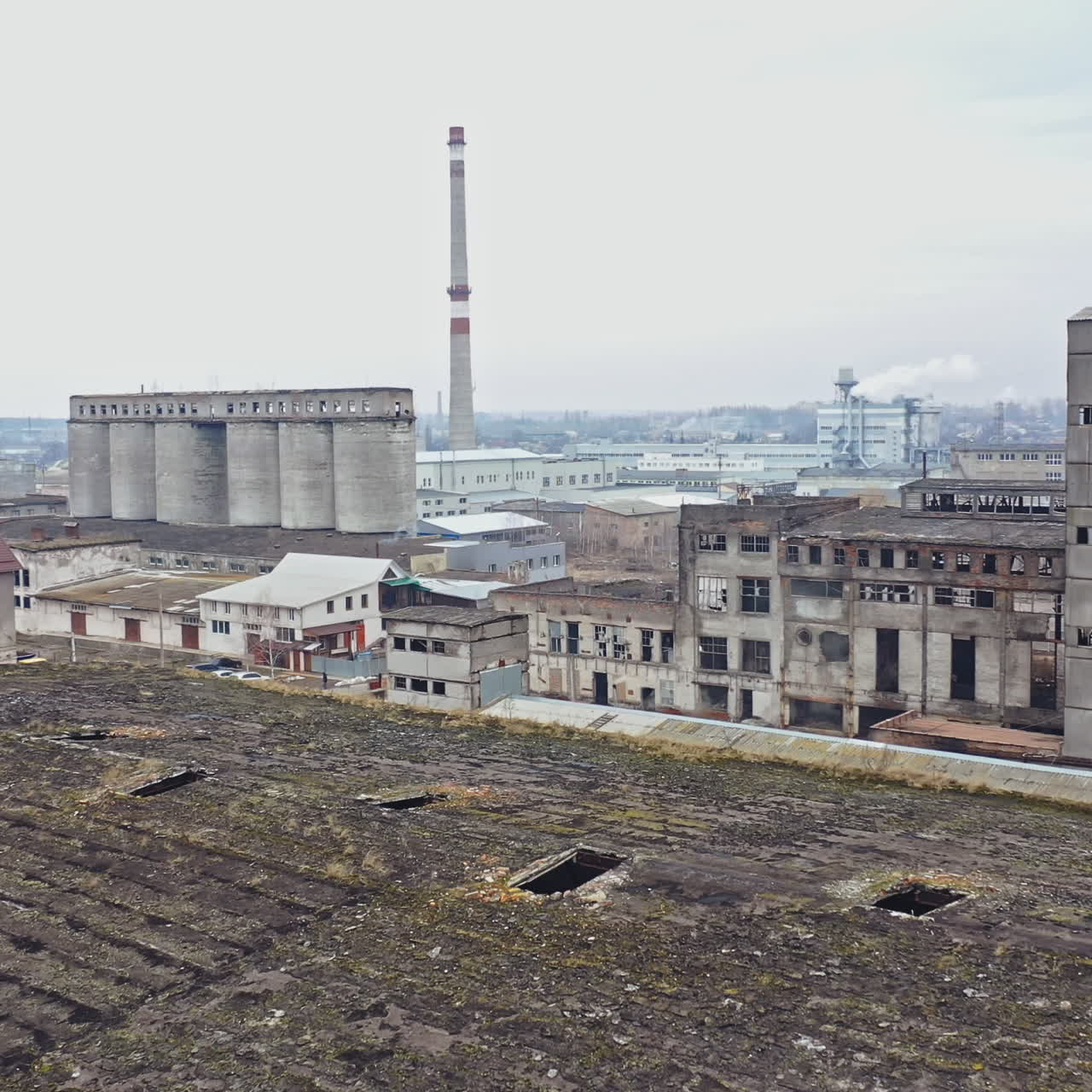 An old abandoned factory hangar with damaged walls on the background of other old structures in the same area. Aerial view.