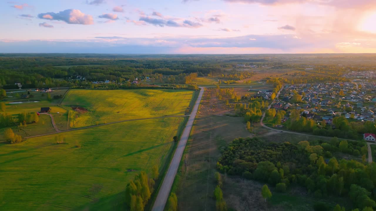 Drone glides above Malutki: straight country road splits verdant fields and leads toward a forest-rimmed horizon in golden evening light. Malutki, Latvia (Malutki, Latvija)