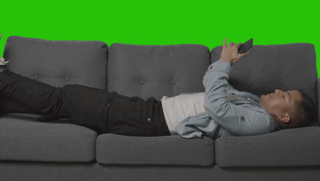 Young Man Lying On Sofa Sneezing And Looking At Mobile Phone Against Green Screen