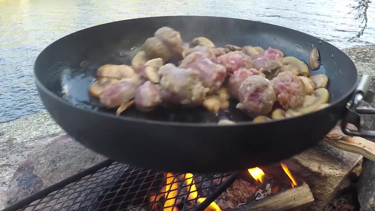 Memasak di atas api terbuka di tepi air. Menggoreng daging dan sayuran di wajan
