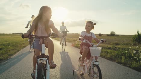 caucasian family of two elementary age girls on foreground and parents in the background riding bikes on village road.