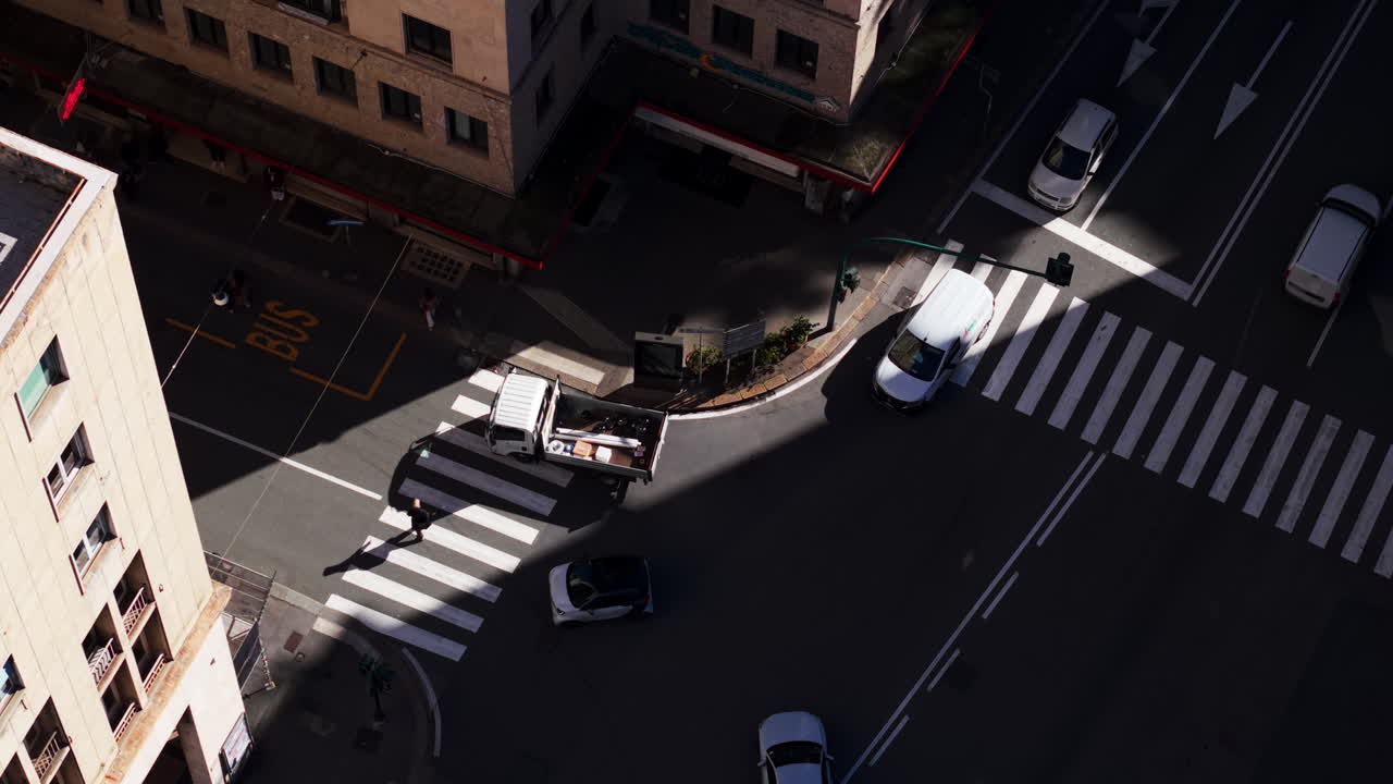 Top-down drone view in Genoa, Liguria, Italy, showing vehicles turning at an intersection as a truck stops to let a pedestrian cross under strong light and shadow contrast