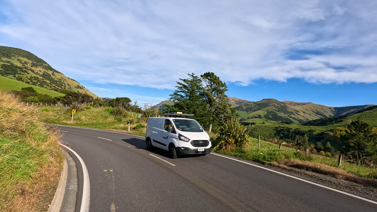 Multiple vehicles travel along a curving rural road surrounded by green hills and mountains under bright daylight, captured with smooth camera panning