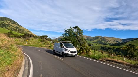 multiple vehicles travel along a curving rural road surrounded by green hills and mountains under bright daylight, captured with smooth camera panning