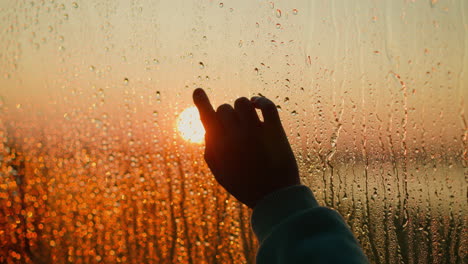 silhouette of finger trying to touch evening sun behind wet window closeup hand touches glass with