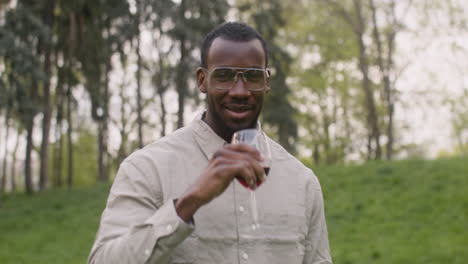 Middle-Aged-Man-Raising-Glass-Of-Wine-While-Looking-At-Camera-In-Park