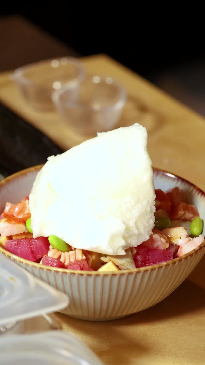 Bidikan close-up seorang koki menambahkan meringue putih ke dalam poke bowl seafood Jepang yang penuh warna.