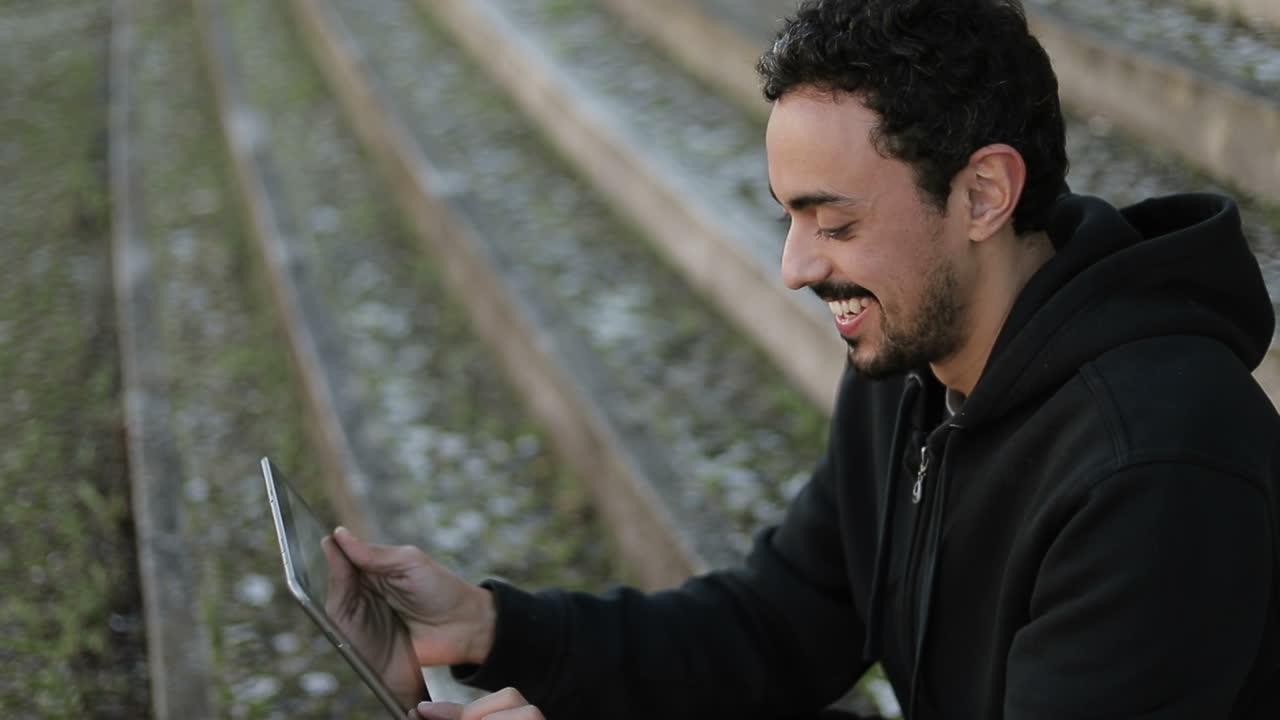 Side view of happy young Arabic handsome man with dark curly hair and beard in black hoodie sitting on stairs outside