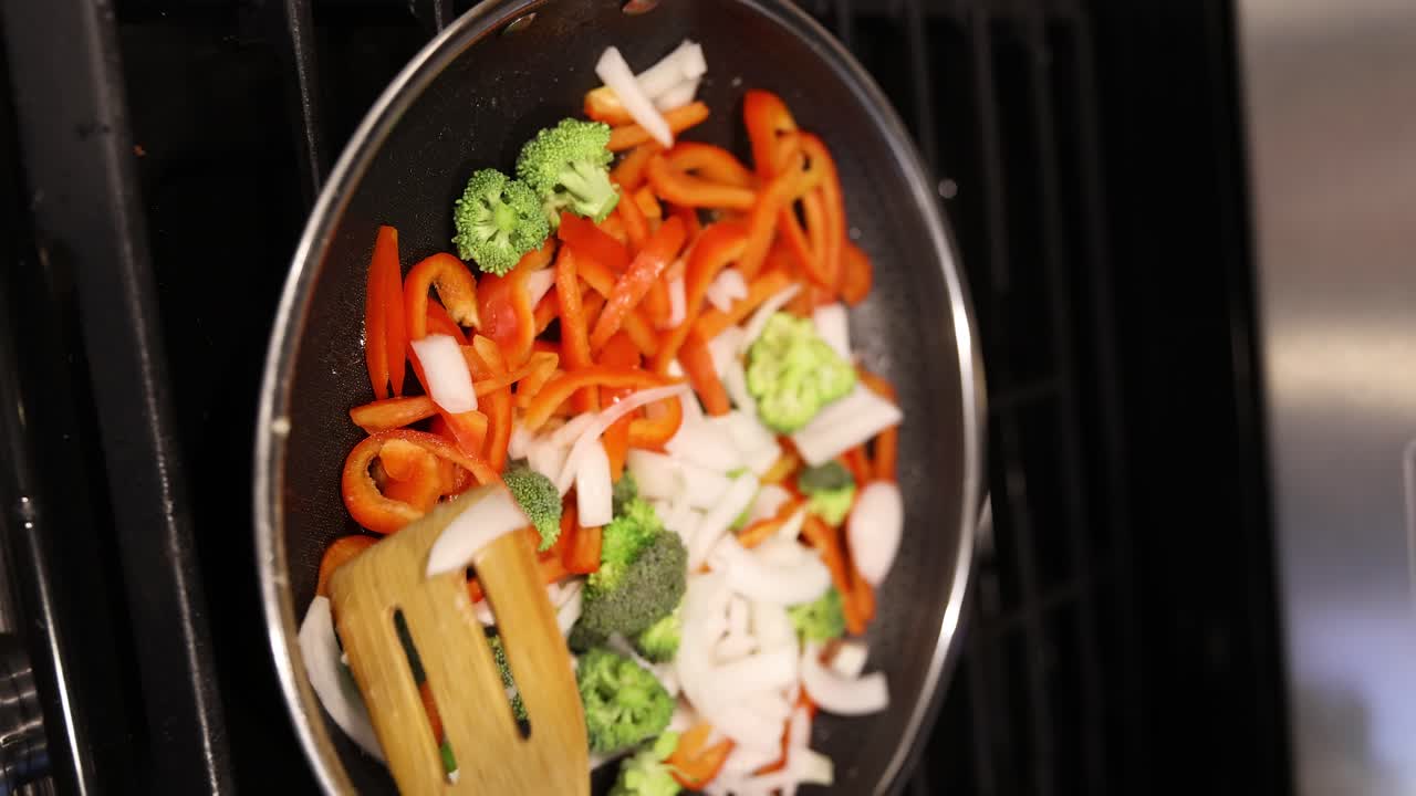 Colorful vegetables sautéed in a pan on a stovetop, being stirred with a wooden spoon