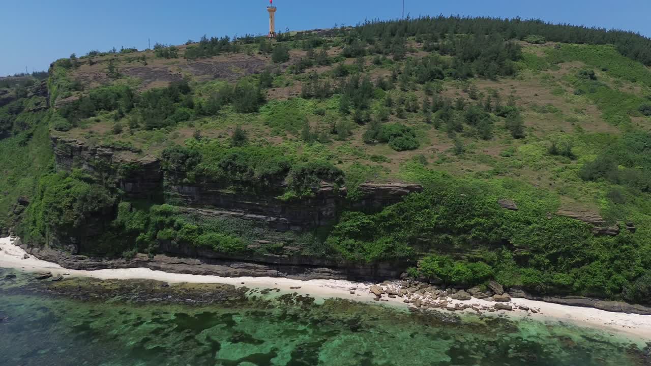 Drone pan left revealing layered cliffs, lush vegetation and turquoise shoreline under bright daylight