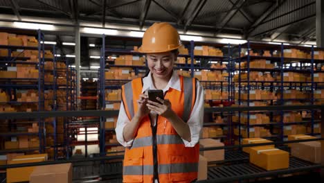 ingeniera asiática con casco de seguridad usando un teléfono inteligente mientras está de pie en el almacén con estantes llenos de productos de entrega