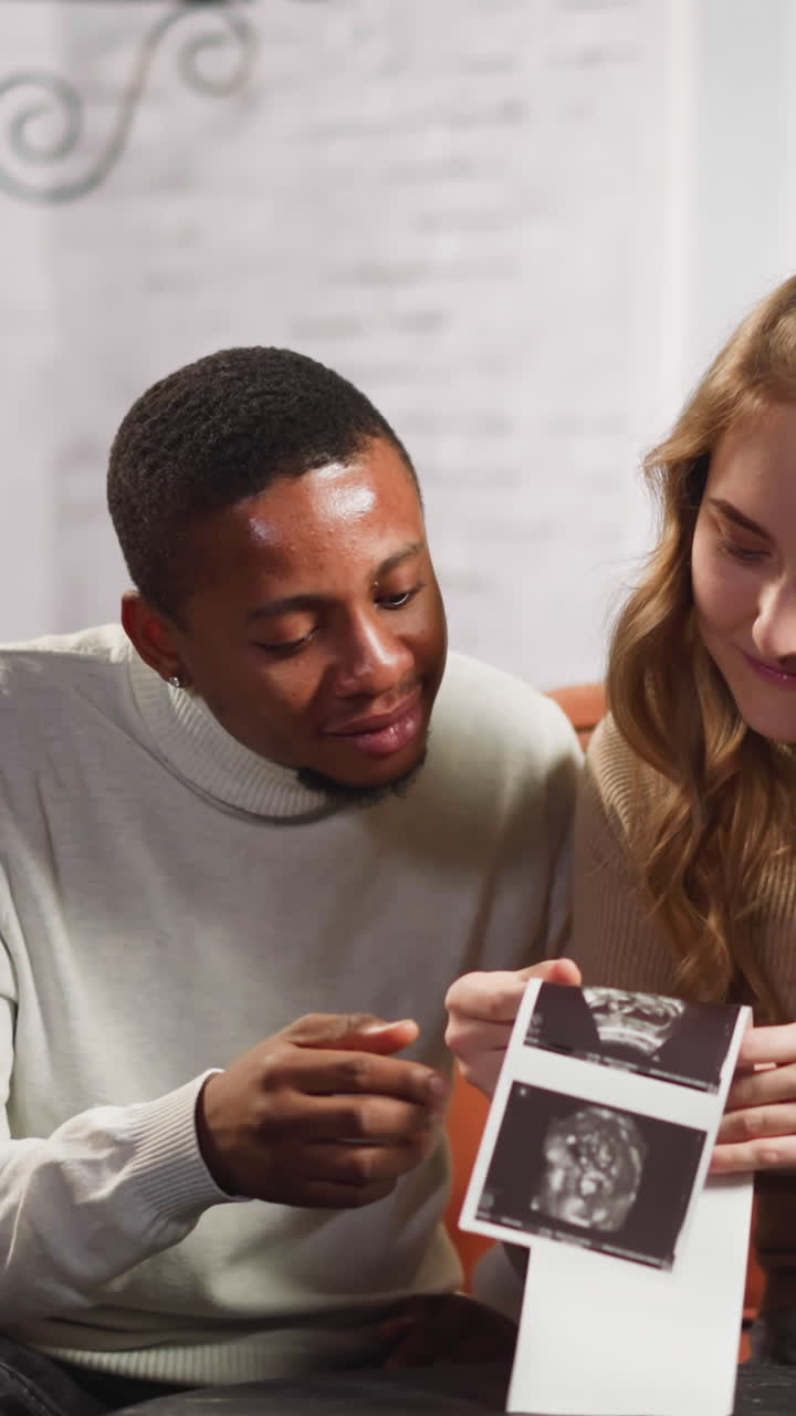 Happy young couple of black man and white woman looks at ultrasonic pictures of expecting baby sitting at small table in living room slow motion