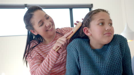 Happy-biracial-mother-brushing-daughter's-hair-in-sunny-bedroom