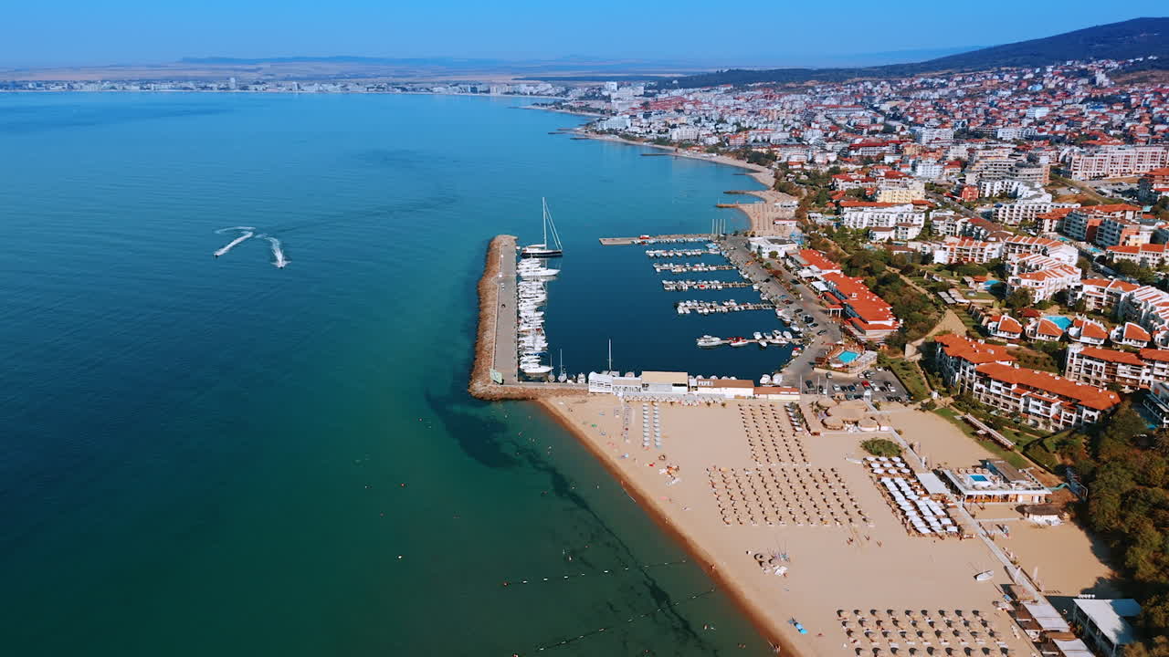 Sunlit sunny beach with deckchairs and umbrellas for vacationers. Multiple hotels dwell on the rocky shore of Bulgaria. Two jet skis ride by the beautiful azure Black Sea