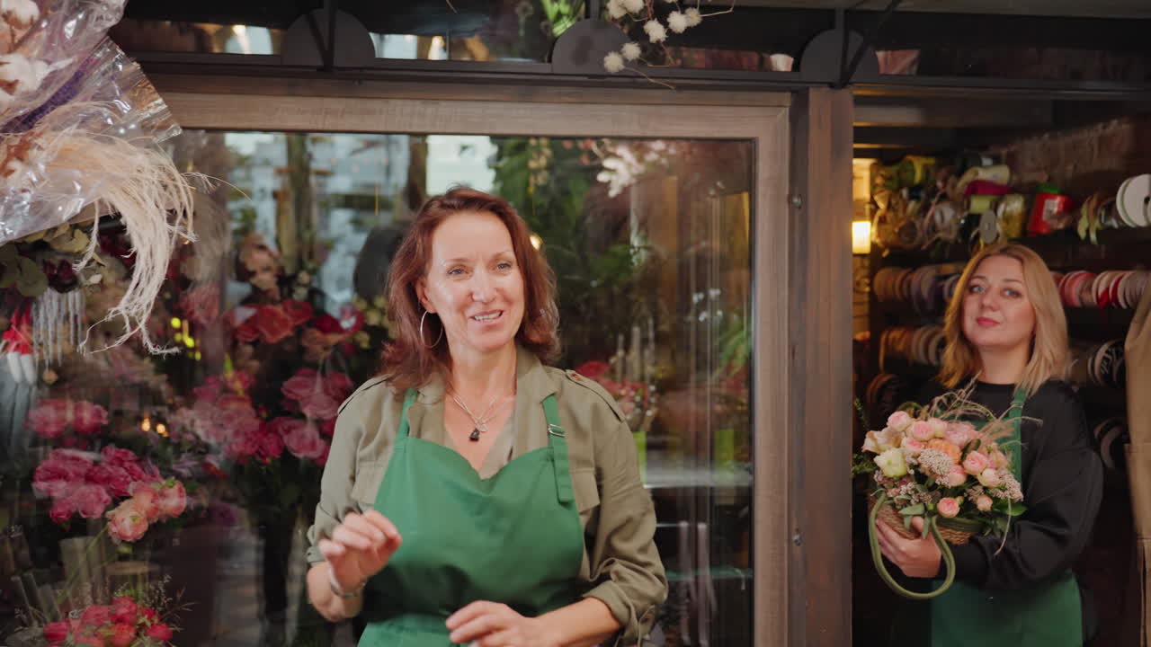 Fiorista che prepara un'esposizione pendente di fiori secchi in una boutique piena di rose e nastri, fiorista (caucasica) concentrata sull'allestimento nuziale, mani che legano steli, grembiule verde, bancone rustico e vasi, collega