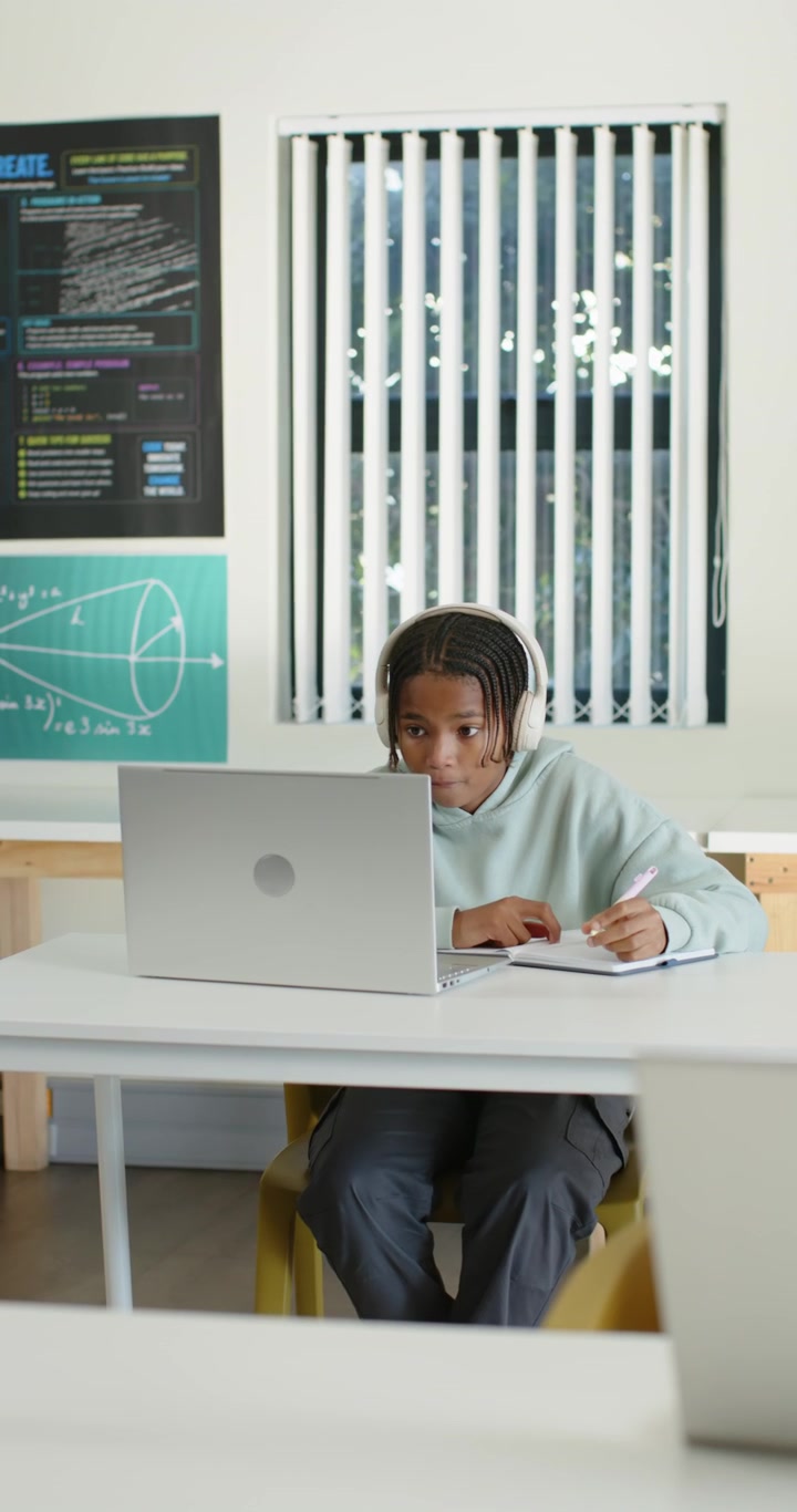 Video verticale: Adolescente afroamericano appoggiato al laptop che prende appunti con le cuffie in classe mentre studia.
