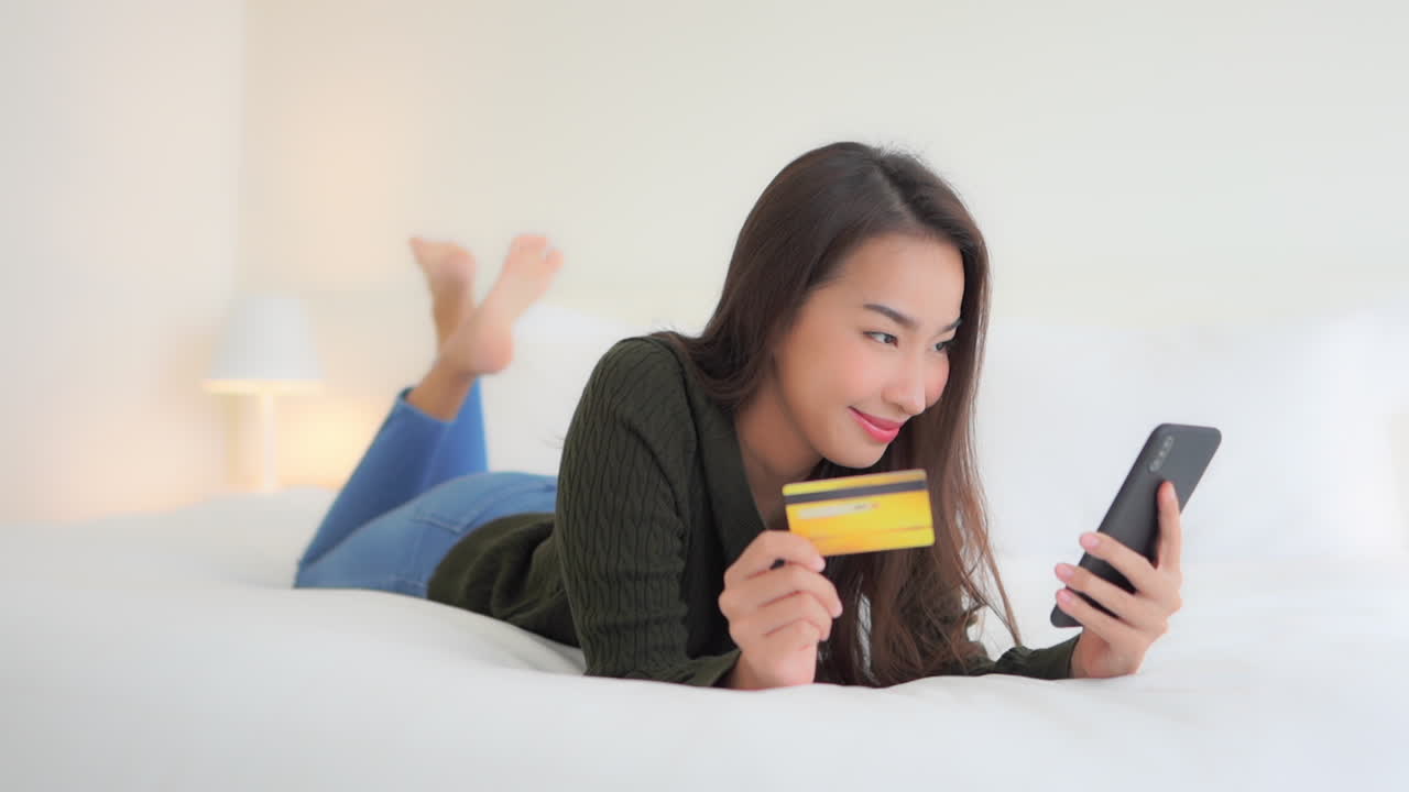 一位美丽的泰国女性躺在豪华特大号床上，用手机输入信用卡号进行在线支付。