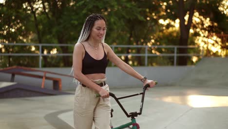 Attractive-stylish-girl-with-dreadlocks-walks-with-bmx-bike-in-skatepark-in-a-recreation-park.-Young-stylish-woman-riding-a-bike