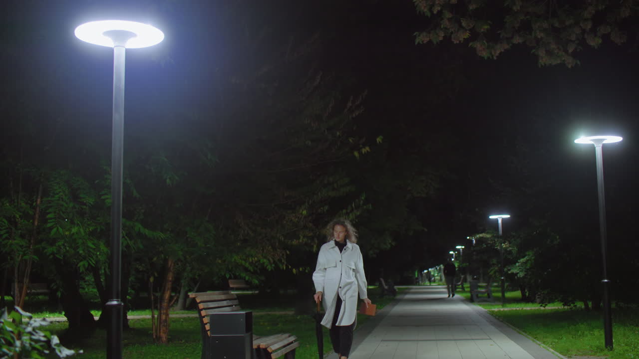 Woman strolls under glowing night streetlights in park, walks toward wooden bench, gently drops umbrella on bench, sits down, crosses legs, opens book to read while others walk in distant background
