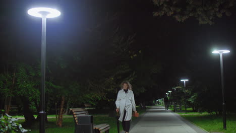 woman strolls under glowing night streetlights in park, walks toward wooden bench, gently drops umbrella on bench, sits down, crosses legs, opens book to read while others walk in distant background
