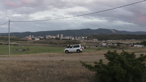 a guy resting on the front hood of a white car parked on the dirt road in tbilisi, georgia on a cloudy day - orbiting drone shot