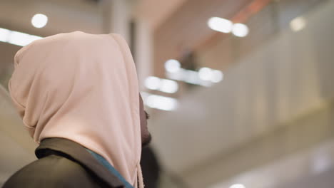 close-up of young man in light hoodie with hood up, standing on escalator in modern shopping mall. his back is partially turned towards the camera, surrounded by bright, sleek mall environment