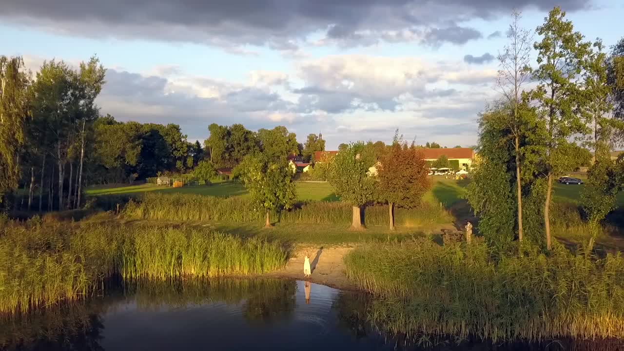 vuelo aéreo por drones de 1 millón de dólares de una mujer rubia solitaria parada sola en el lago reflejando el atardecer de verano en un lago en un pequeño pueblo en brandeburgo, alemania