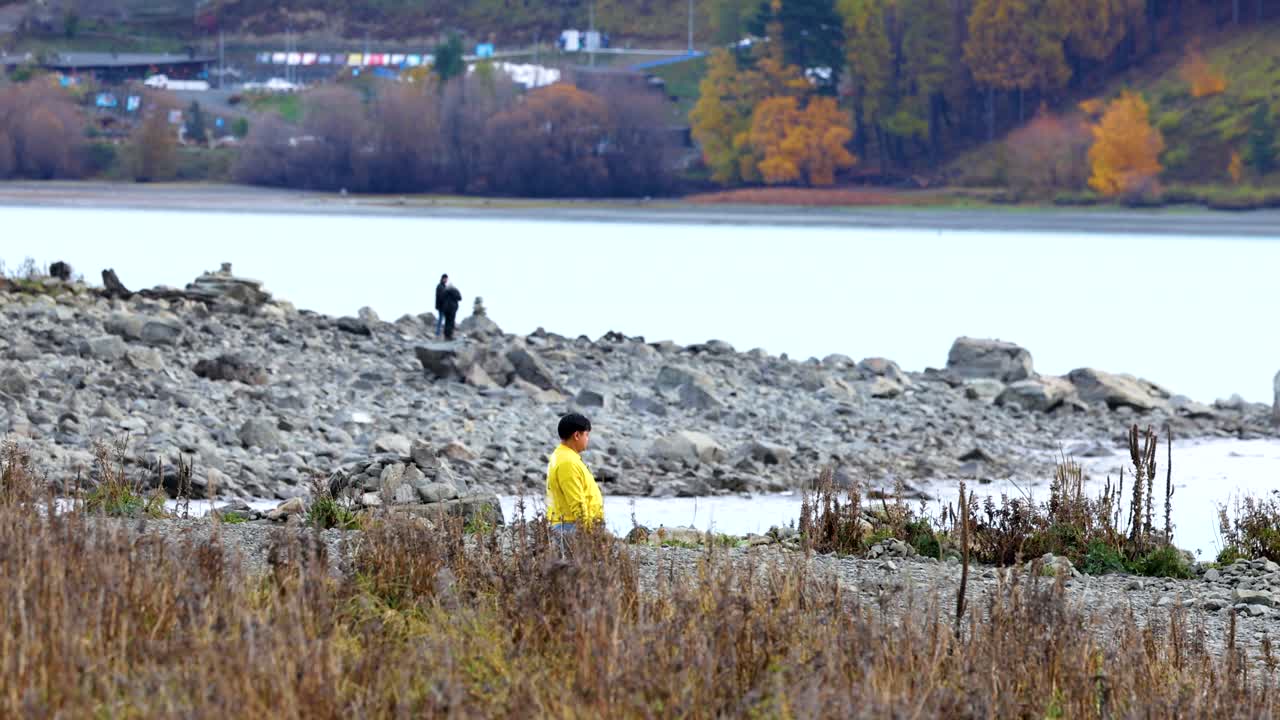 A person in a yellow jacket walks along Lake Tekapo's rocky shore, surrounded by autumn foliage under soft lighting