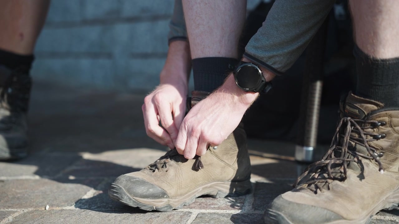 Primo piano delle mani di un escursionista che allaccia i lacci di scarponi da trekking usurati su una pavimentazione in pietra. Si prepara per un'avventura di trekking in montagna all'aperto con uno smartwatch al polso