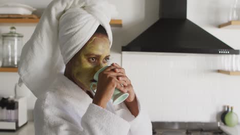 Happy-african-american-woman-with-beauty-mask-on-face,-drinking-coffee-in-kitchen