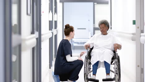 mujer médica diversa hablando con una mujer anciana en silla de ruedas en el pasillo del hospital, cámara lenta