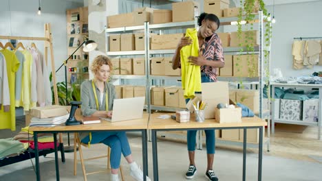 diseñadora afroamericana empacando ropa ordenada en cajas y hablando por teléfono mientras el gerente de ventas caucásico escribe en una laptop en una tienda de ropa de moda