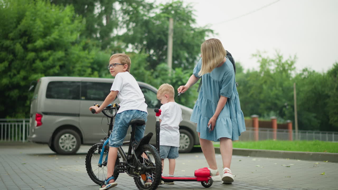 A mother bends while combing her younger son's hair, who looks quiet and thoughtful, the older sibling stands beside them with a bicycle, while a red scooter is nearby
