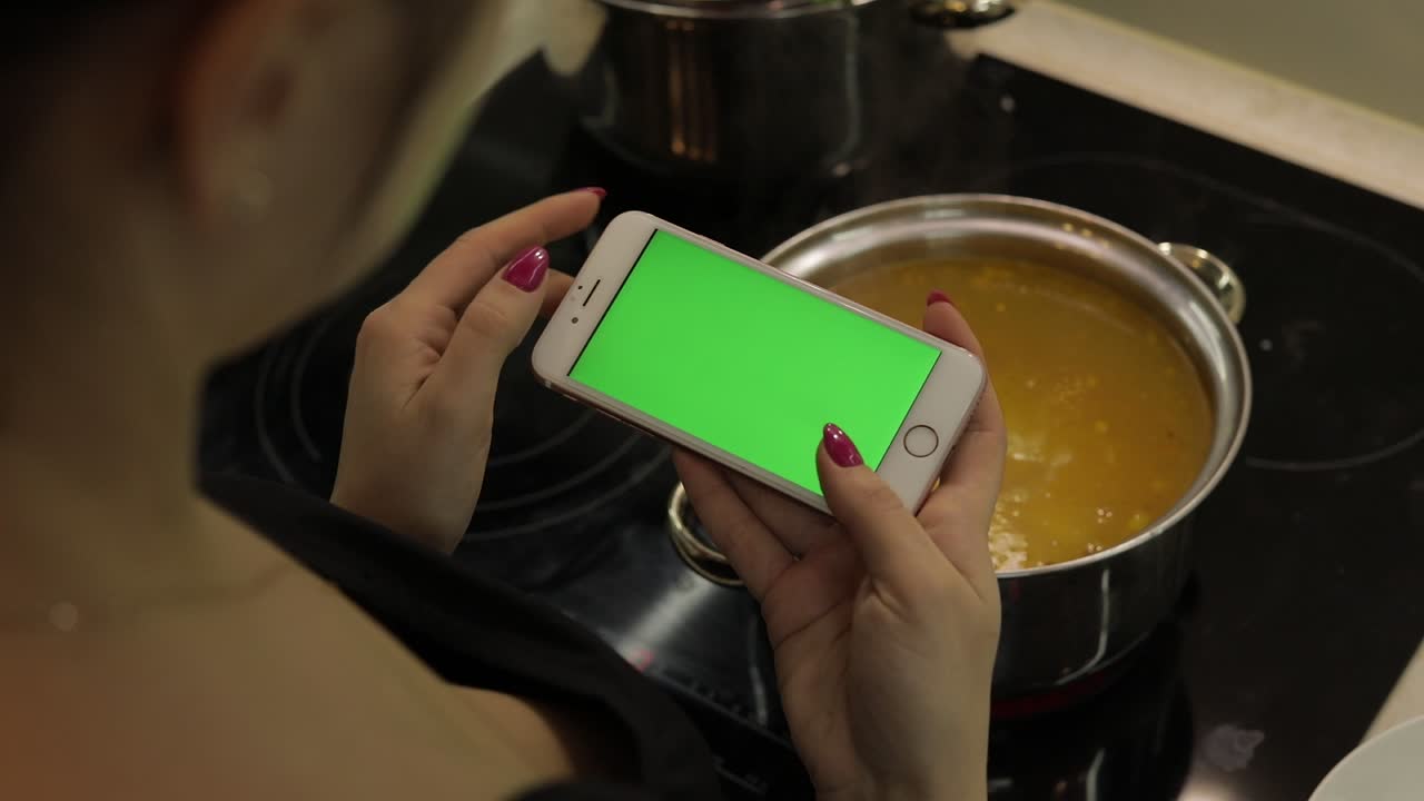 mujer está sosteniendo un teléfono inteligente con una pantalla verde, y cocinando sopa