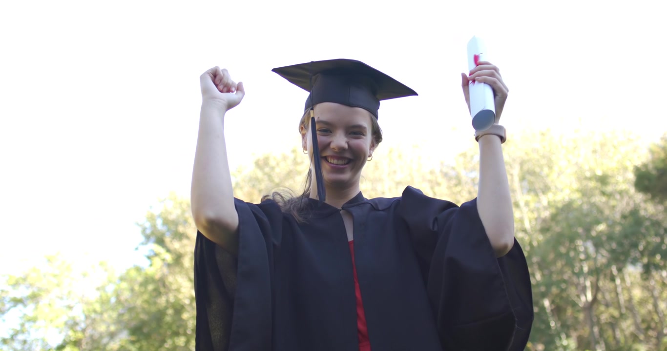 Laureata che festeggia la laurea nel parco, tenendo in mano il diploma e il tocco e la toga, alzando le braccia.