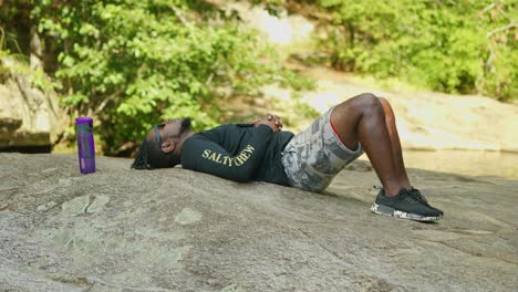 a man lays down and relaxes on a giant rock in the middle of the woods on a bright sunny day