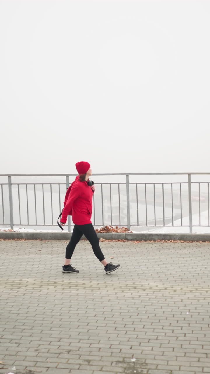 Side view of woman walking on patterned pathway with bag over shoulder near iron railing in foggy atmosphere with distant cars below bridge and dry foliage scattered on snowy ground