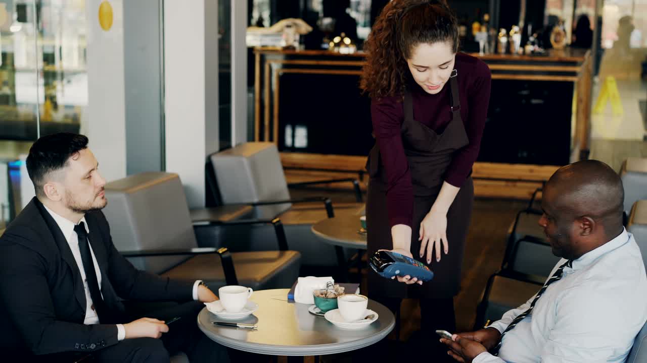dos hombres de negocios multirraciales con ropa formal pagan la factura en línea usando un teléfono inteligente en un café moderno. una joven camarera de pie y manteniendo una terminal de efectivo portátil.
